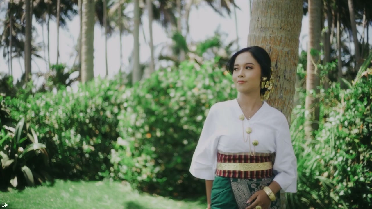Pasangan berbusana adat Bali berjalan bergandengan di atas rumput menuju pantai berpohon kelapa. — 1