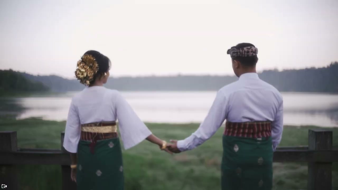 Pasangan berbusana adat Bali berjalan bergandengan di atas rumput menuju pantai berpohon kelapa. — 3
