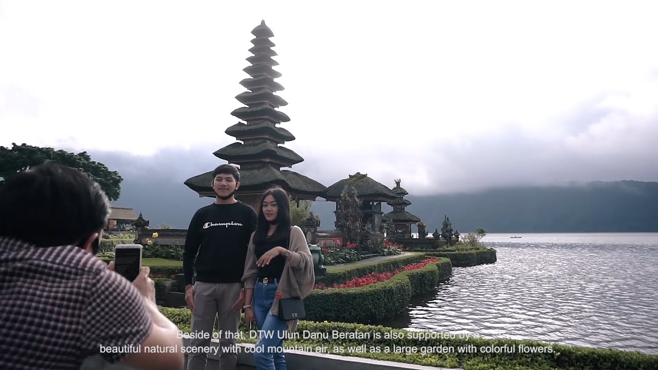 Sepasang pengunjung berfoto di depan meru bertingkat di tepi Danau Beratan saat langit mendung, dengan seorang pria berkemeja kotak-kotak memotret mereka memakai ponsel dari depan.