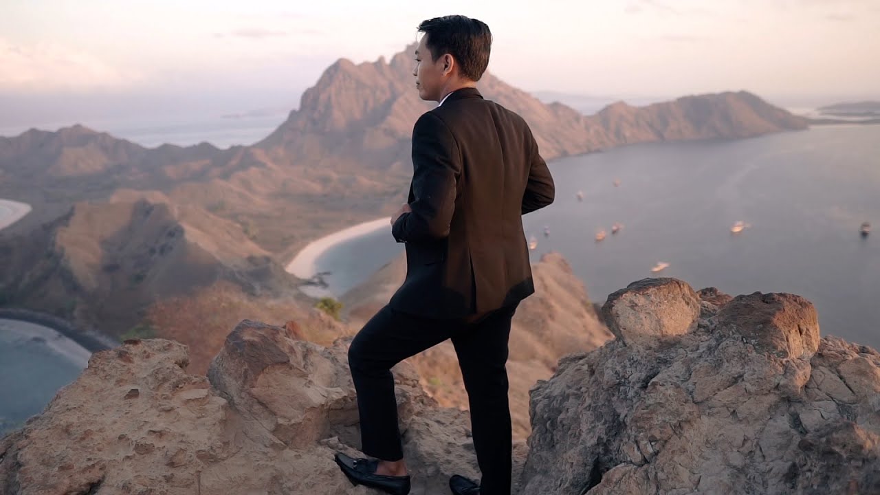 Pria berjas gelap berdiri membelakangi kamera di punggung bukit berbatu, memandang teluk berpasir putih dan perahu-perahu kecil saat cahaya rendah.