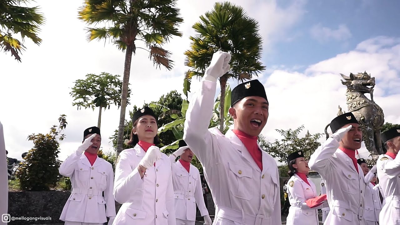 Anggota pasukan pengibar bendera berseragam putih berkerah merah, berpeci hitam, dan bersarung tangan putih; satu orang mengepalkan tangan ke atas sambil berteriak dan yang lain memberi hormat di bawah langit biru berawan.