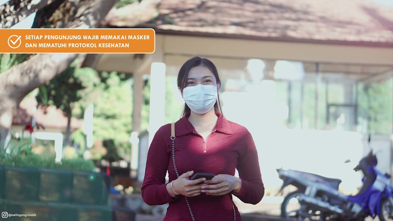 Perempuan berbaju merah marun bermasker memegang ponsel di depan bangunan beratap genteng, dengan spanduk oranye berisi imbauan memakai masker di sudut kiri atas.