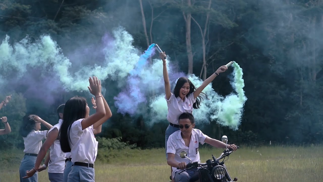 Siswa berseragam putih bersorak di lapangan rumput sementara seorang siswa berkacamata hitam mengendarai motor klasik dan seorang siswi berdiri di boncengan mengangkat dua flare asap biru dan hijau.