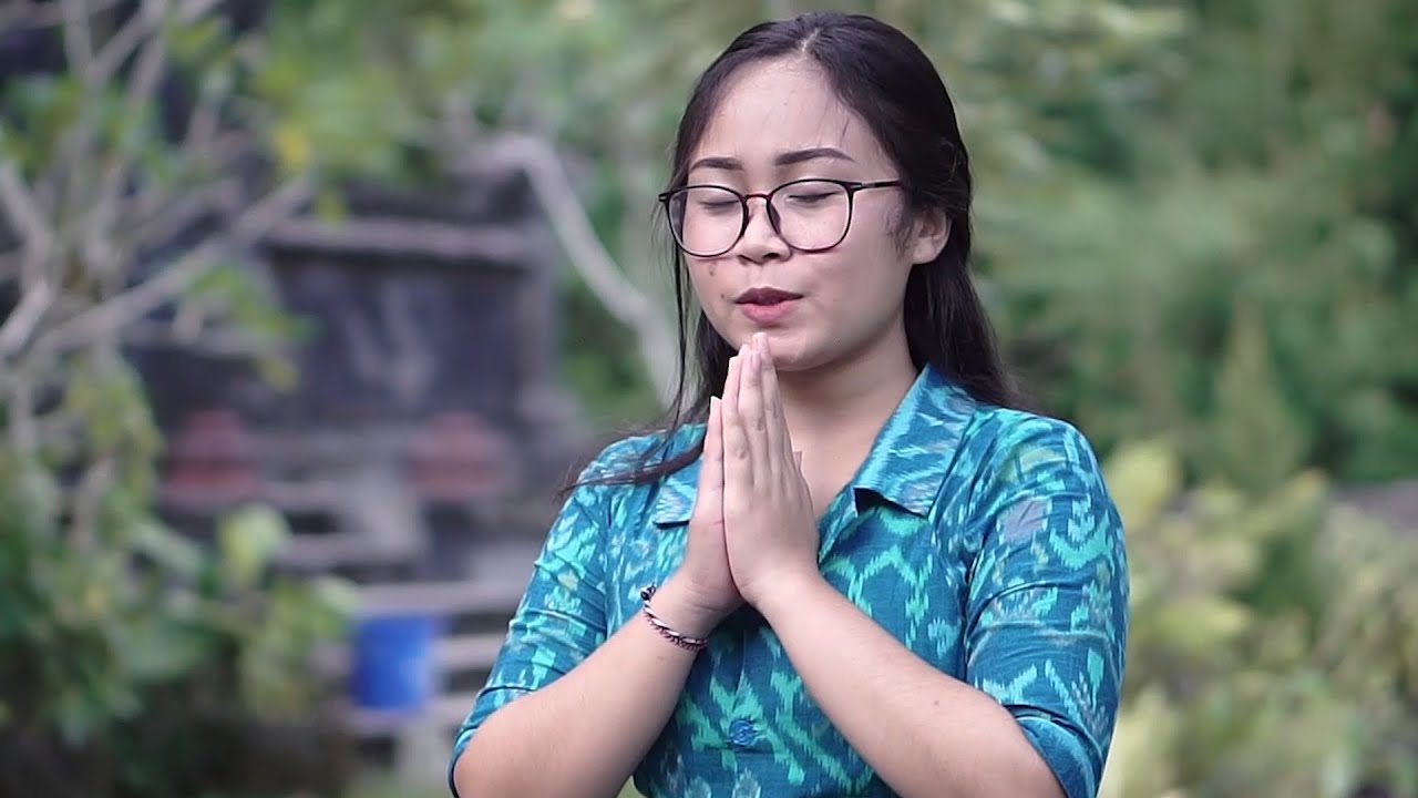 Potret dekat perempuan muda berkacamata dan berkemeja bermotif biru kehijauan, mata terpejam dengan kedua telapak tangan dikatupkan di depan dada, berlatar dedaunan dan pelinggih yang buram.