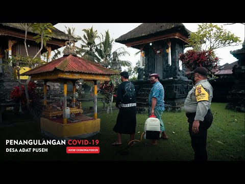 Tiga orang, termasuk seorang polisi dan seorang pria berpakaian adat Bali yang menggendong alat semprot punggung, berdiri di halaman pura menjelang malam dengan teks penanggulangan COVID-19 Desa Adat Puseh di sudut kiri bawah.