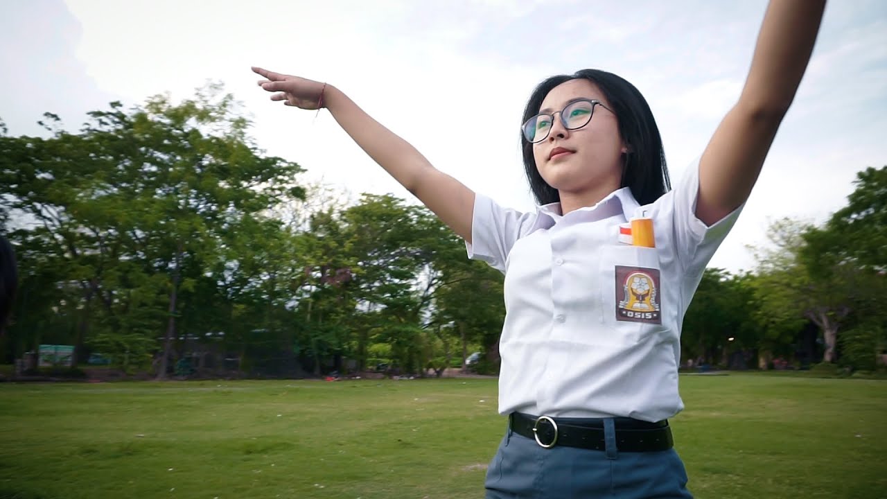 Siswi berseragam putih dengan lencana OSIS di saku merentangkan kedua tangan di lapangan rumput dengan barisan pohon dan langit terang di belakangnya.