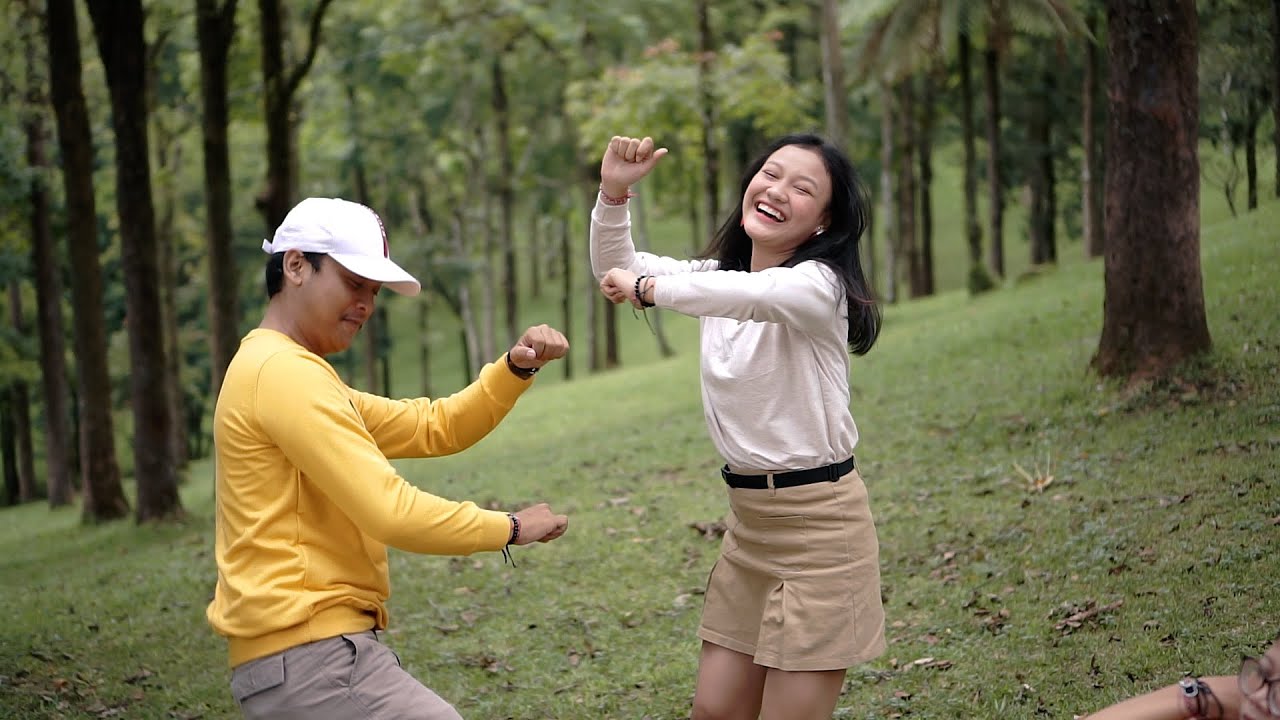 Pria bersweter kuning dan bertopi putih menari berhadapan dengan perempuan muda berkaus krem yang tertawa di lereng rumput di antara pepohonan tinggi.