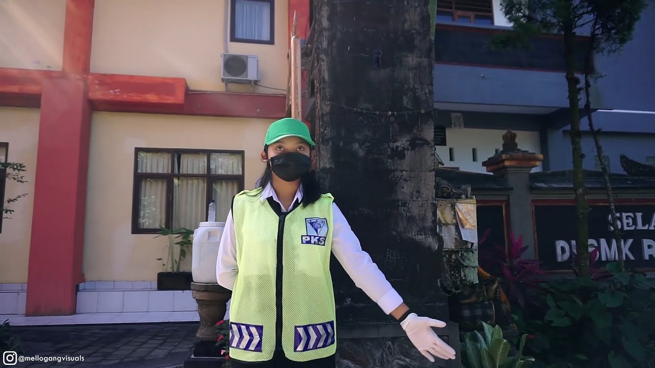 Siswi bertopi hijau, bermasker hitam, dan berompi PKS kuning menyala merentangkan satu tangan di depan gedung sekolah berdinding krem dan merah.